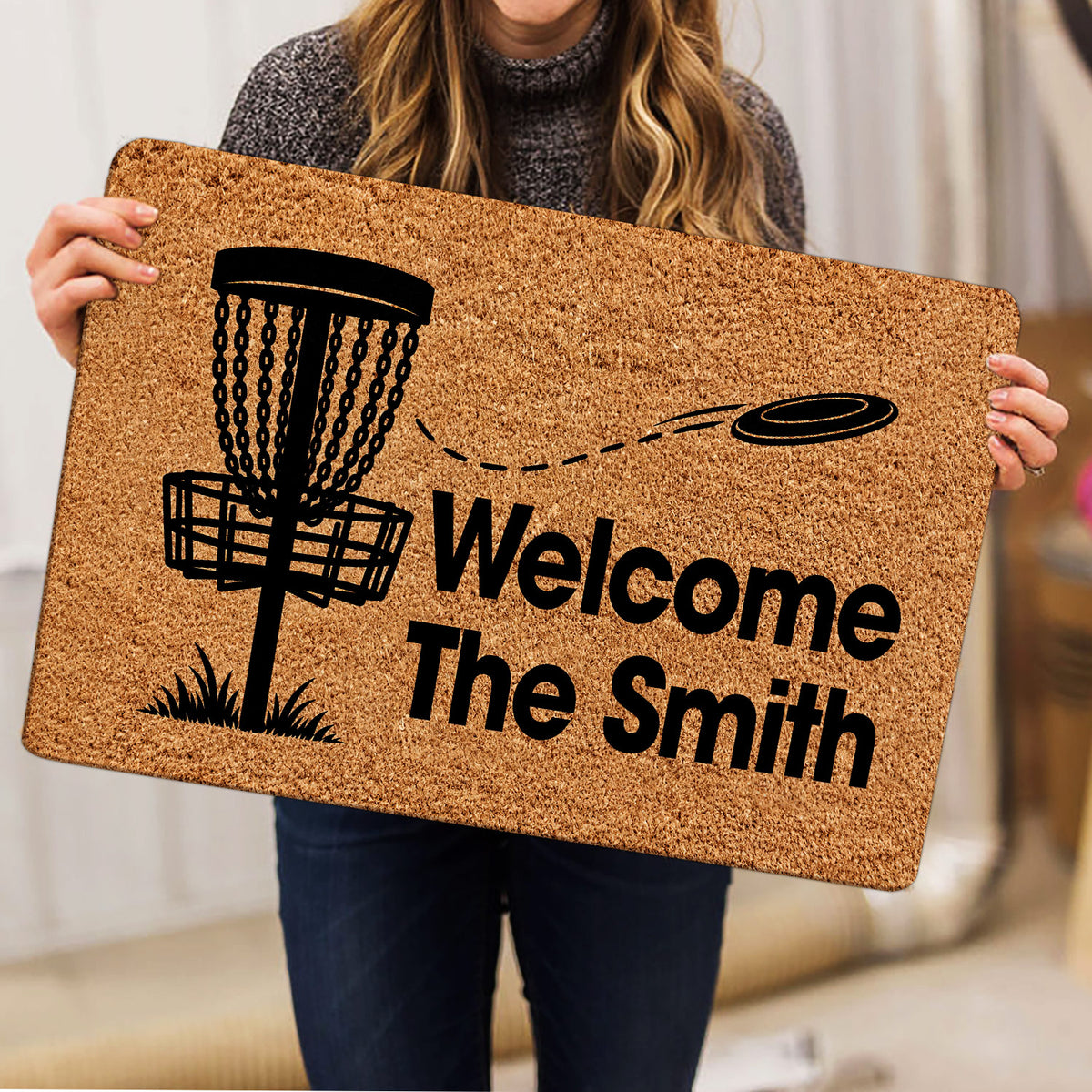 Person holding a disc golf doormat featuring a basket, flying disc, and bold welcome artwork