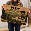 Person holding a disc golf doormat featuring a basket, mountains, and evergreen trees