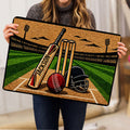 Cricket doormat held indoors, showing detailed bat, wickets, helmet, ball and green pitch artwork