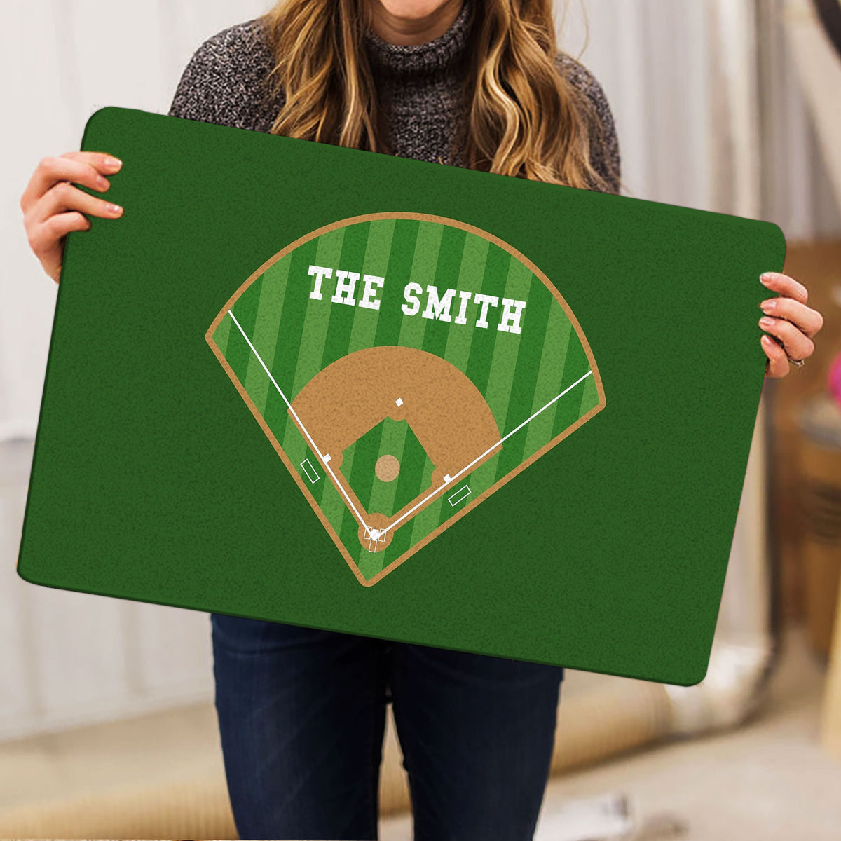 Person holding a green baseball doormat showing a ballpark diamond and striped field artwork