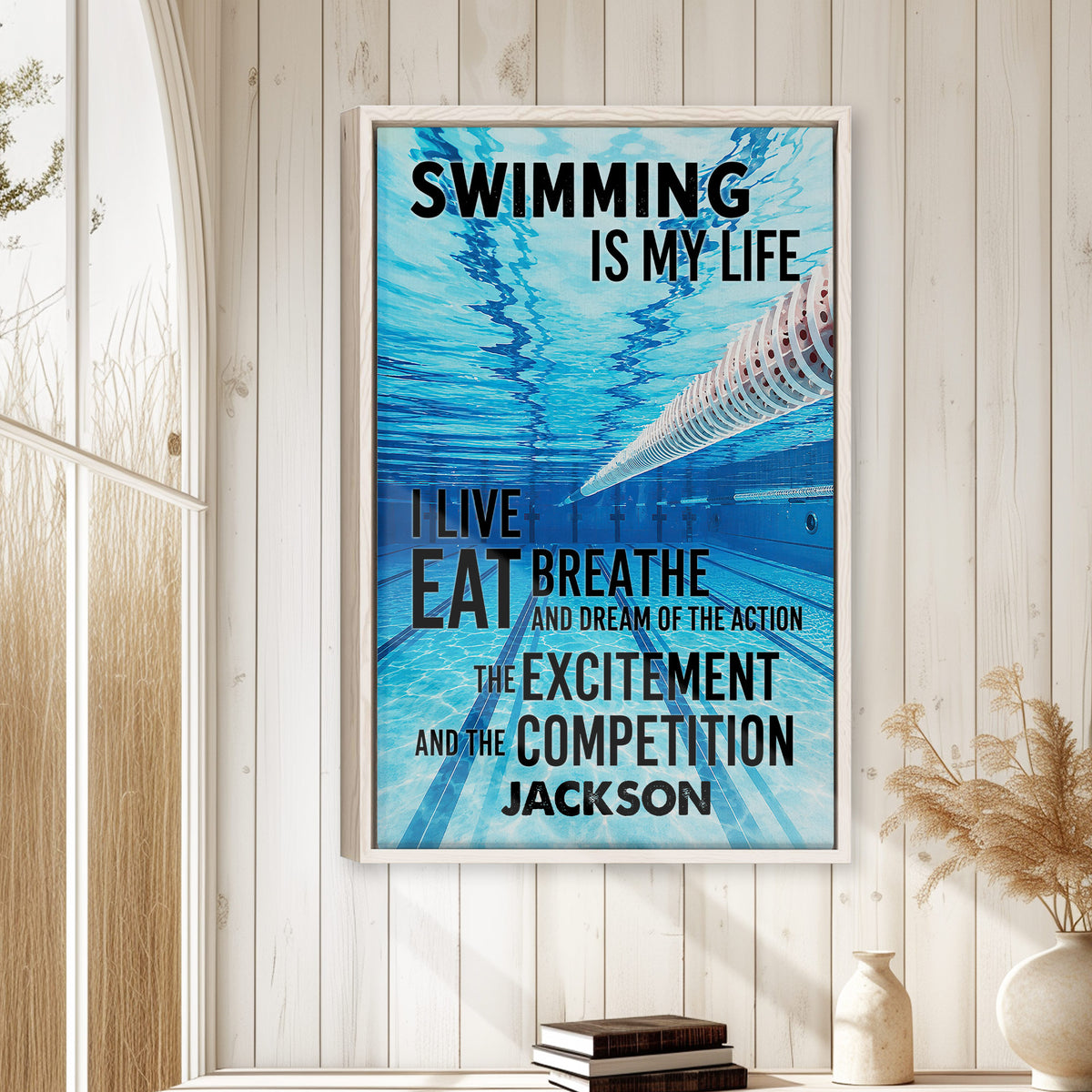 Swimming wall art displayed in a bright room on a white wood-paneled wall above stacked books