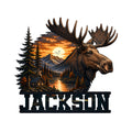 Moose metal sign featuring a large antlered moose, mountain lake, pine forest, cabin, and sunset scene