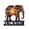 Elephant-shaped metal sign featuring an African savanna sunset, acacia trees, mountains, and a family herd