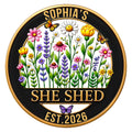 Round She Shed metal sign with colorful wildflowers, butterflies, and a gold-trimmed black design
