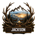 Deer metal sign with large antlers framing two deer, mountain peaks, pine trees, a lake, and a rustic cabin