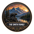 Round bear metal sign featuring a black bear overlooking a mountain lake at sunset