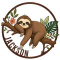 Round sloth metal sign featuring a smiling sloth lounging on a branch with tropical leaves and flowers