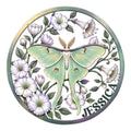 Round metal sign featuring a mint green moth surrounded by delicate lavender flowers and leafy botanical details