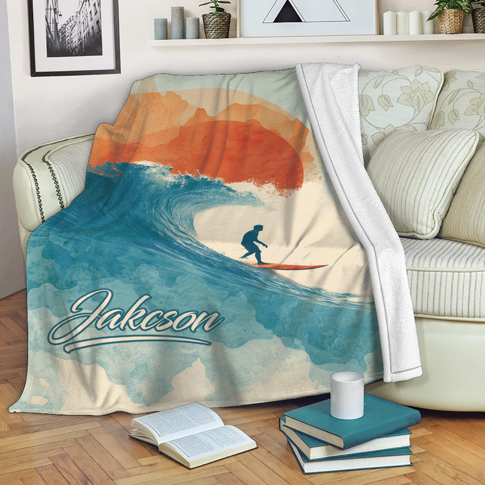 Custom surfing blanket draped over a sofa with a surfer, curling wave, and warm watercolor sunset design