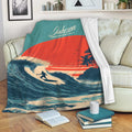 Surfing throw blanket displayed on a sofa with ocean waves, sunset colors, and a surfer silhouette