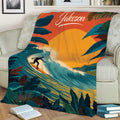 Surfing throw blanket displayed on a couch with dramatic waves, tropical foliage, and a warm orange sunset