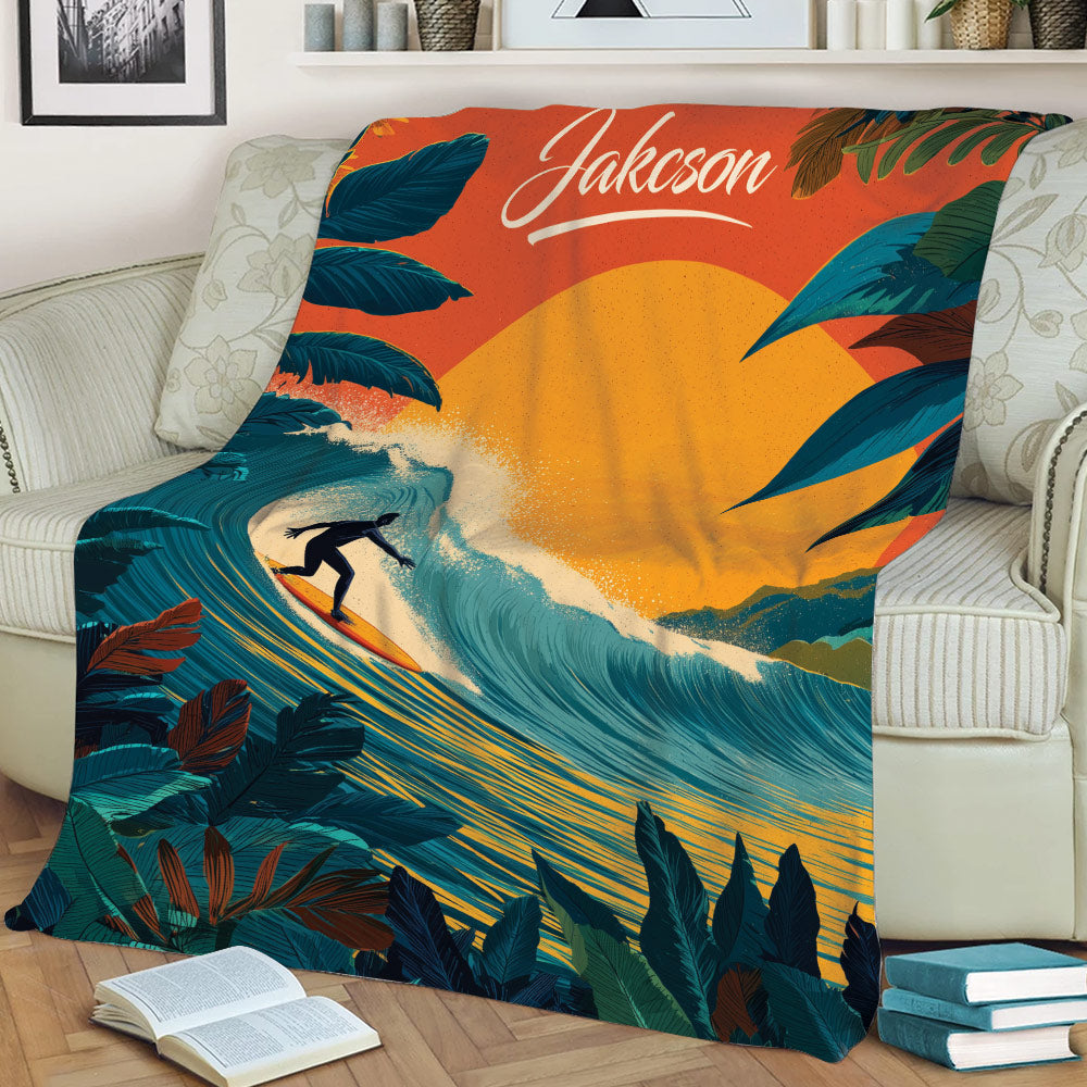 Surfing throw blanket displayed on a couch with dramatic waves, tropical foliage, and a warm orange sunset