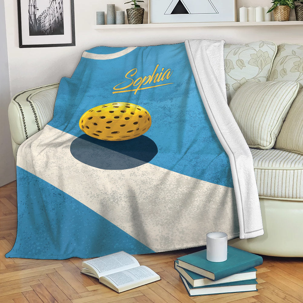 Custom blue pickleball blanket draped over a sofa with yellow ball artwork and a cozy living room setting