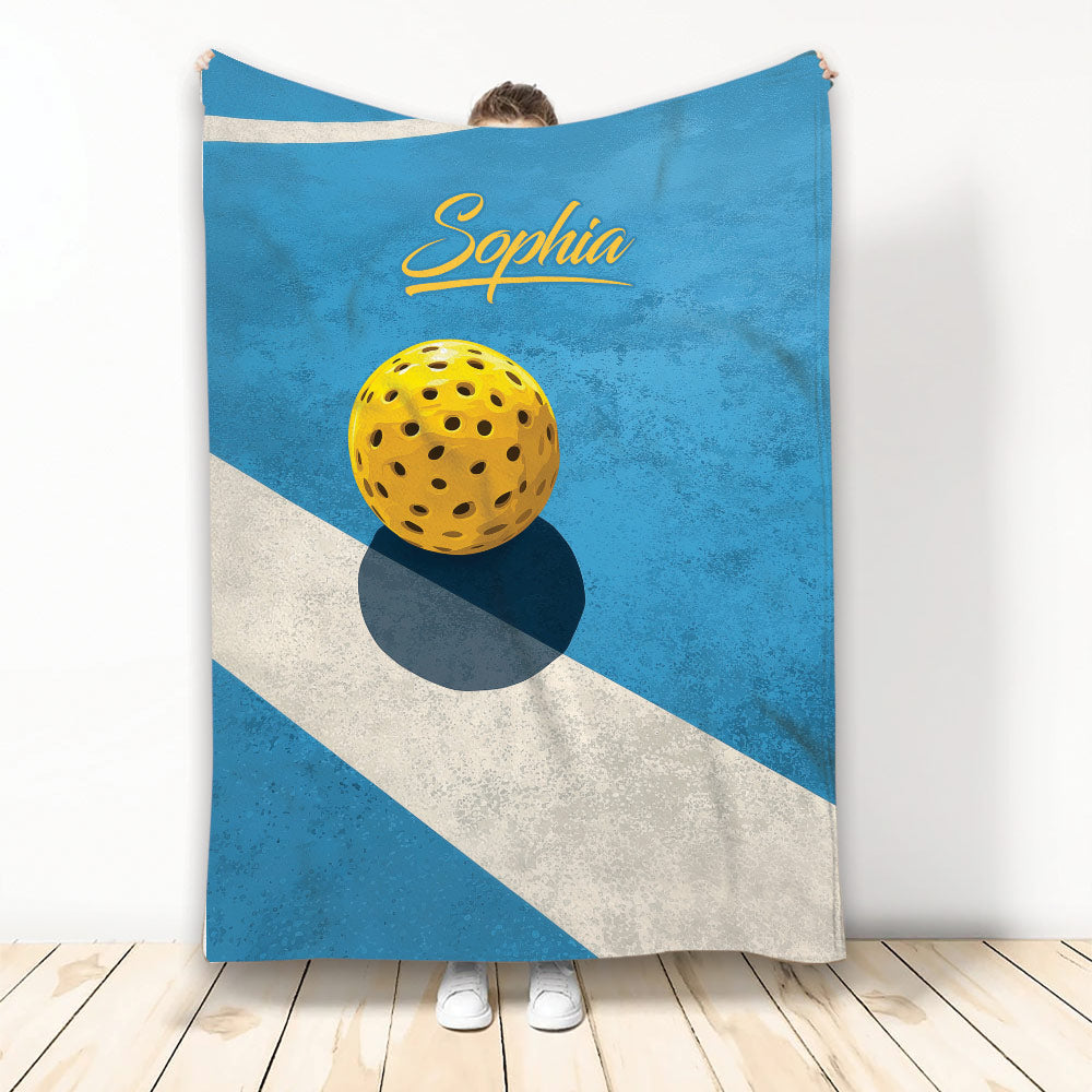 Blue pickleball throw blanket displayed with a yellow perforated ball and bold geometric court-inspired artwork