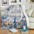 Hanukkah throw blanket displayed on a sofa with blue floral menorah artwork, dreidels, gifts, and holiday treats