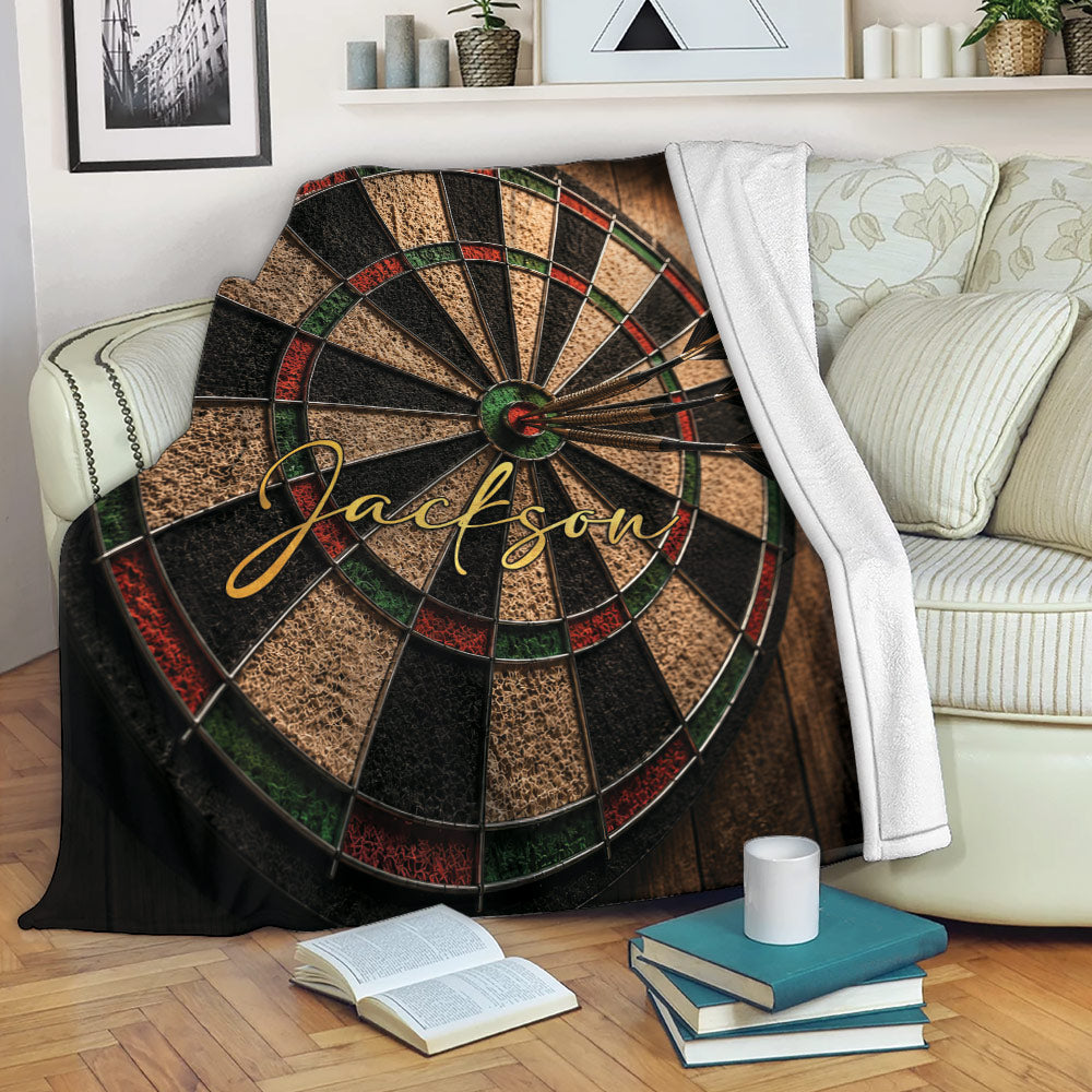 Dart board throw blanket draped over a sofa, featuring darts and rustic black, red, green, and tan artwork