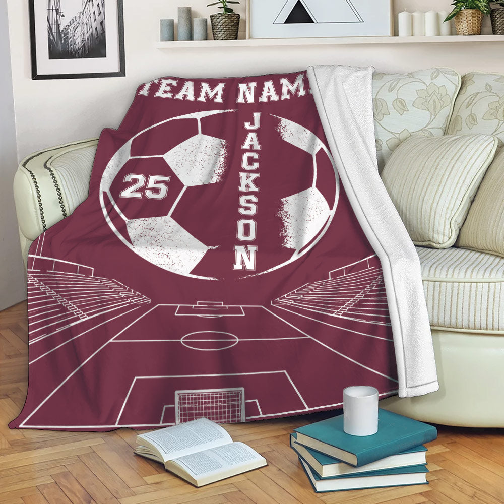 Maroon soccer throw blanket draped over a sofa with a large ball, stadium scene and field design