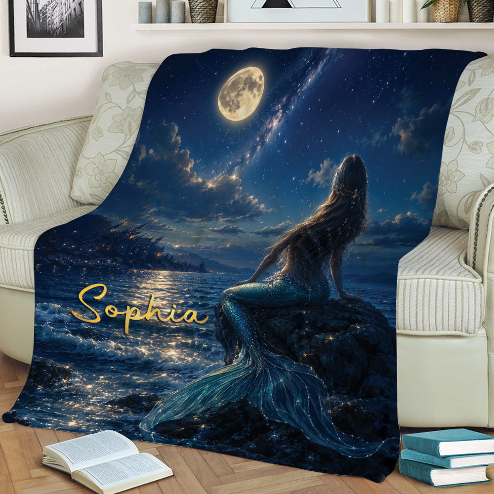 Mermaid throw blanket displayed on a sofa with a moonlit sea, starry sky, rocky shore, and blue tail