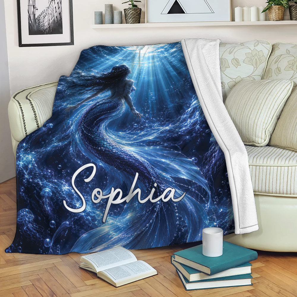 Mermaid throw blanket draped over a sofa with deep blue underwater artwork, sea life, bubbles, and radiant light rays