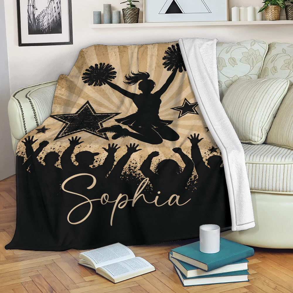 Cheerleading blanket displayed on a sofa with a jumping cheerleader, pom-poms, stars, and crowd silhouettes