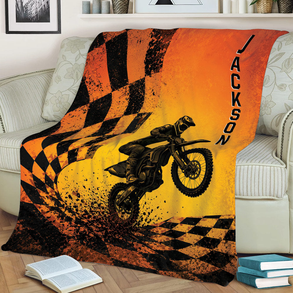 Orange and black motocross throw blanket displayed on a couch with dirt bike action artwork and racing flag graphics