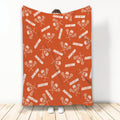 Orange lacrosse blanket displayed upright with repeating sticks, helmets, balls, and sports artwork