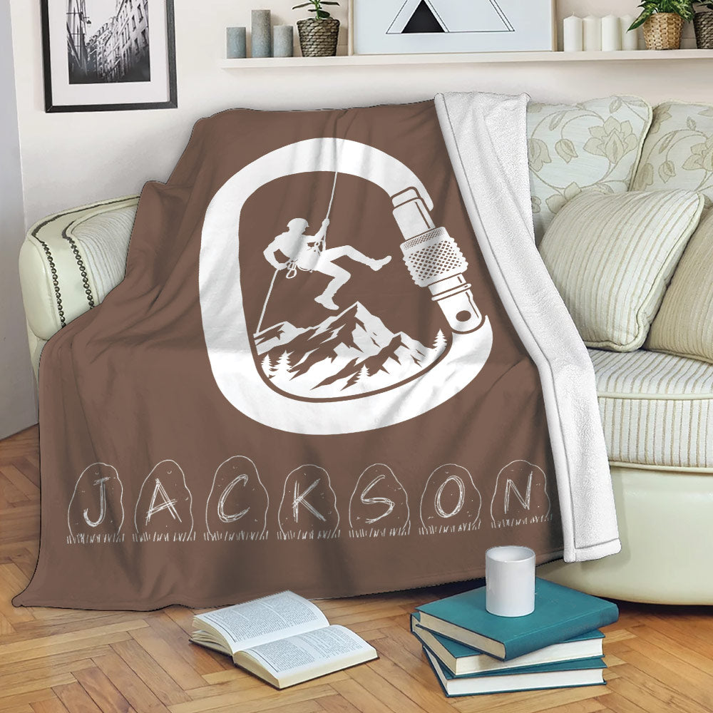 Brown rock climbing throw blanket draped over a sofa with climber, mountain, rope, and carabiner artwork