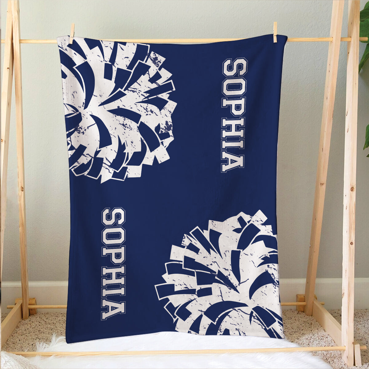 Blue cheerleading blanket hanging on a wooden display frame with large white pom-pom designs