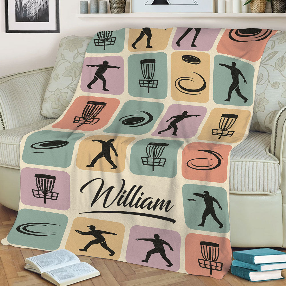 Disc Golf blanket displayed on a couch with colorful panels, disc golf baskets, discs, and player silhouettes