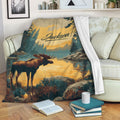 Moose blanket draped over a sofa, featuring a rustic lakeside forest scene in gold and teal tones
