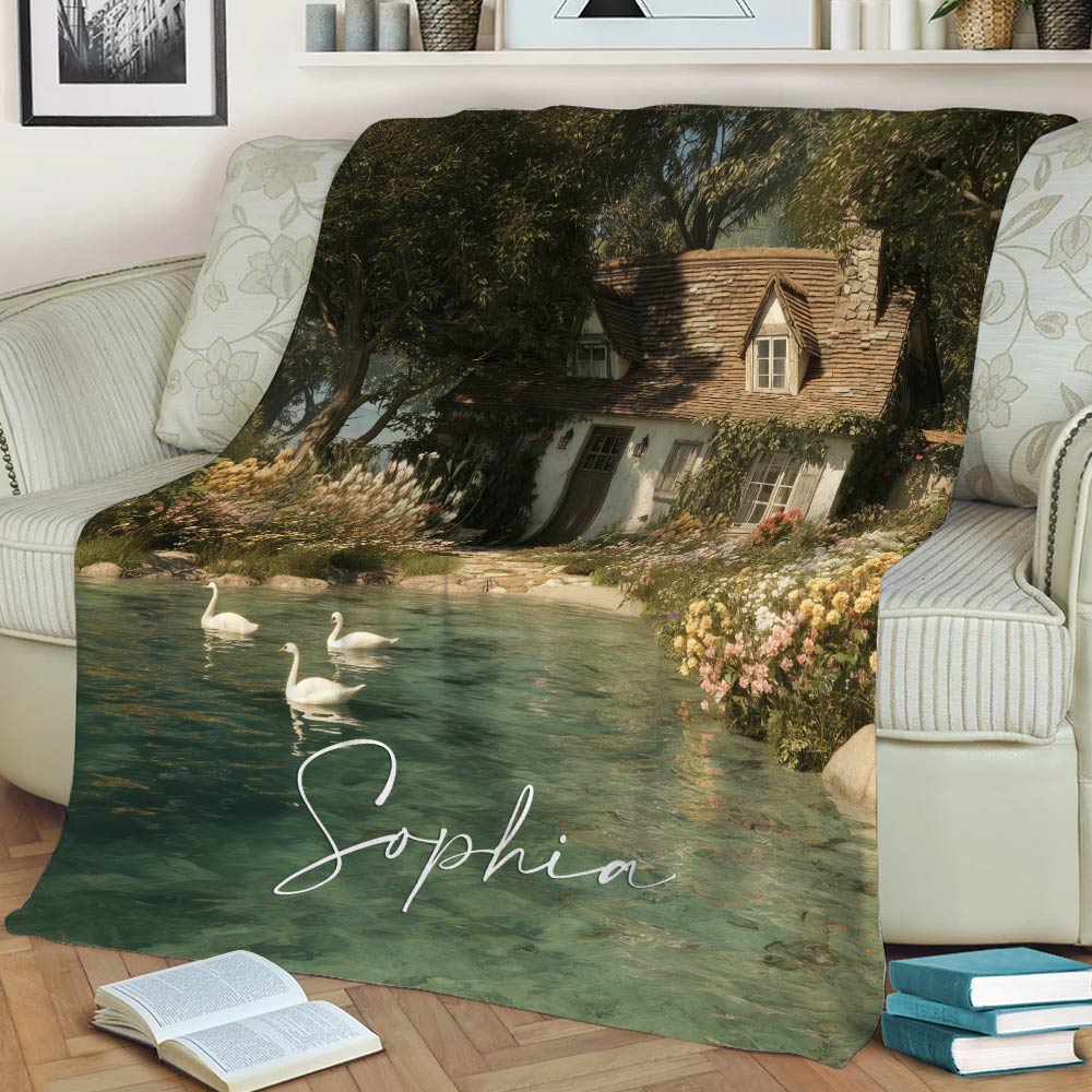 Lake house blanket displayed across a sofa, featuring a cottage beside a flower-lined pond with graceful swans