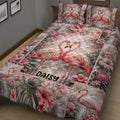 Flamingo quilt bedding displayed across a bed with pink hibiscus flowers and tropical foliage