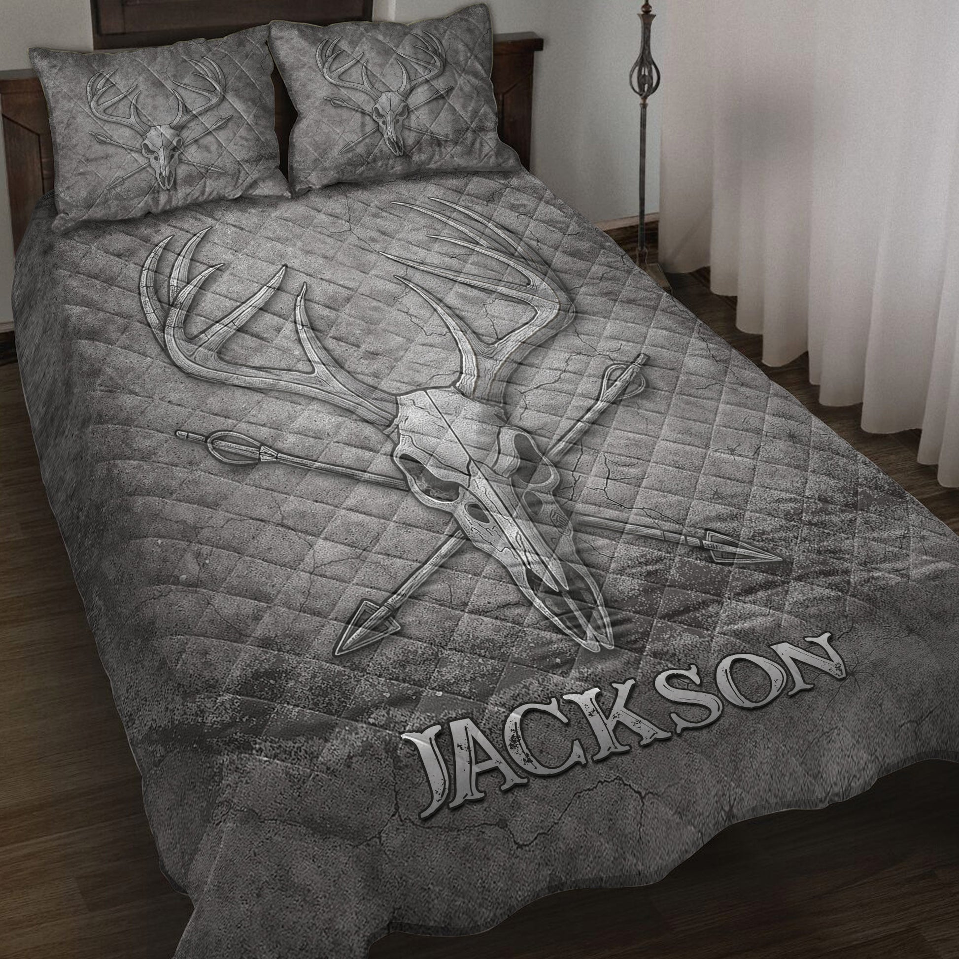 Gray hunting quilt blanket displayed on a bed with deer skull, antler, and crossed arrow artwork