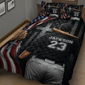 Close bedroom view of a baseball quilt featuring a ballplayer from behind, bat, glove, stars, and red white and blue artwork