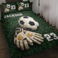 Soccer quilt blanket displayed on a bed with goalkeeper gloves, soccer ball artwork, and green field design