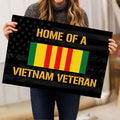 Person holding a Vietnam Veteran doormat featuring patriotic stars, gold lettering, and a red yellow green flag