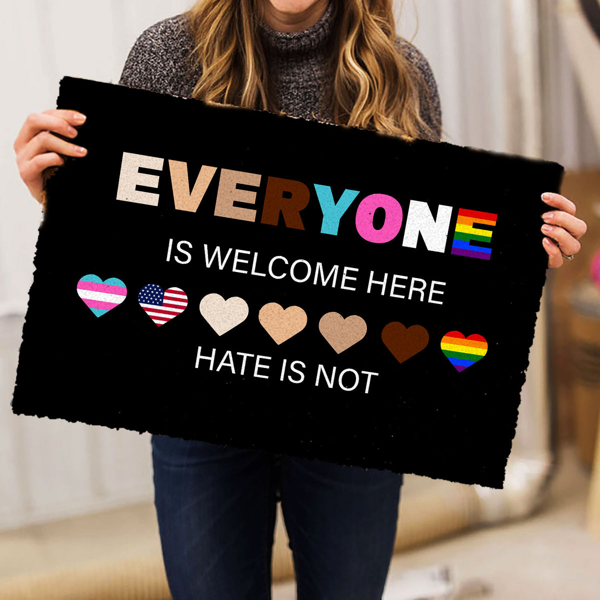 Person holding a black LGBT doormat with colorful hearts and inclusive wording