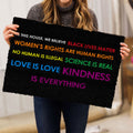 Person holding a black LGBT doormat with colorful lettering and an inclusive message