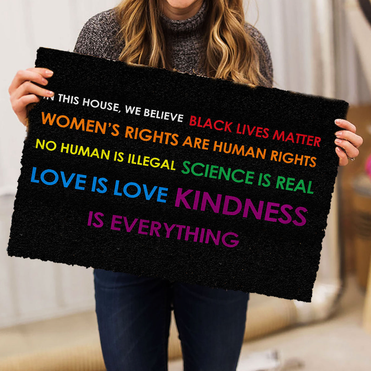 Person holding a black LGBT doormat with colorful lettering and an inclusive message