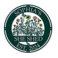 Round She Shed metal sign with colorful wildflowers, butterflies, and a deep green decorative border